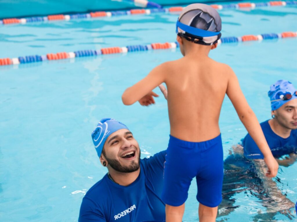 Un infante entrando a una piscina, a punto de tomar clases de natación para niños que ya saben nadar, con la ayuda de un maestro