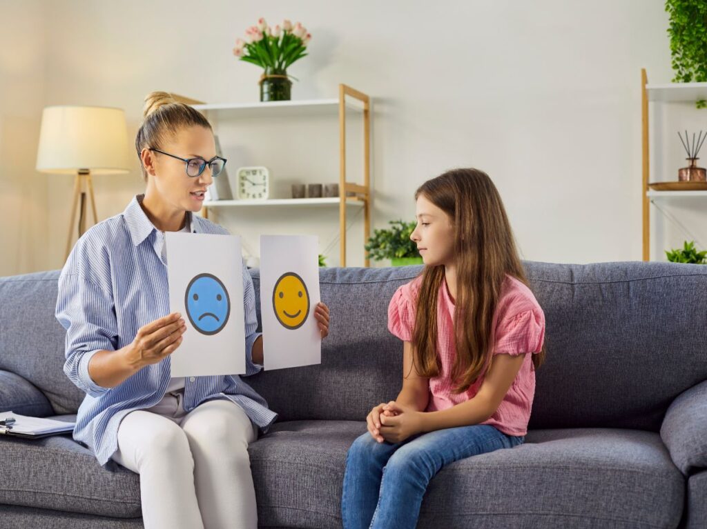 Una niña con su maestra quien le muestra dos caritas, una feliz y una triste